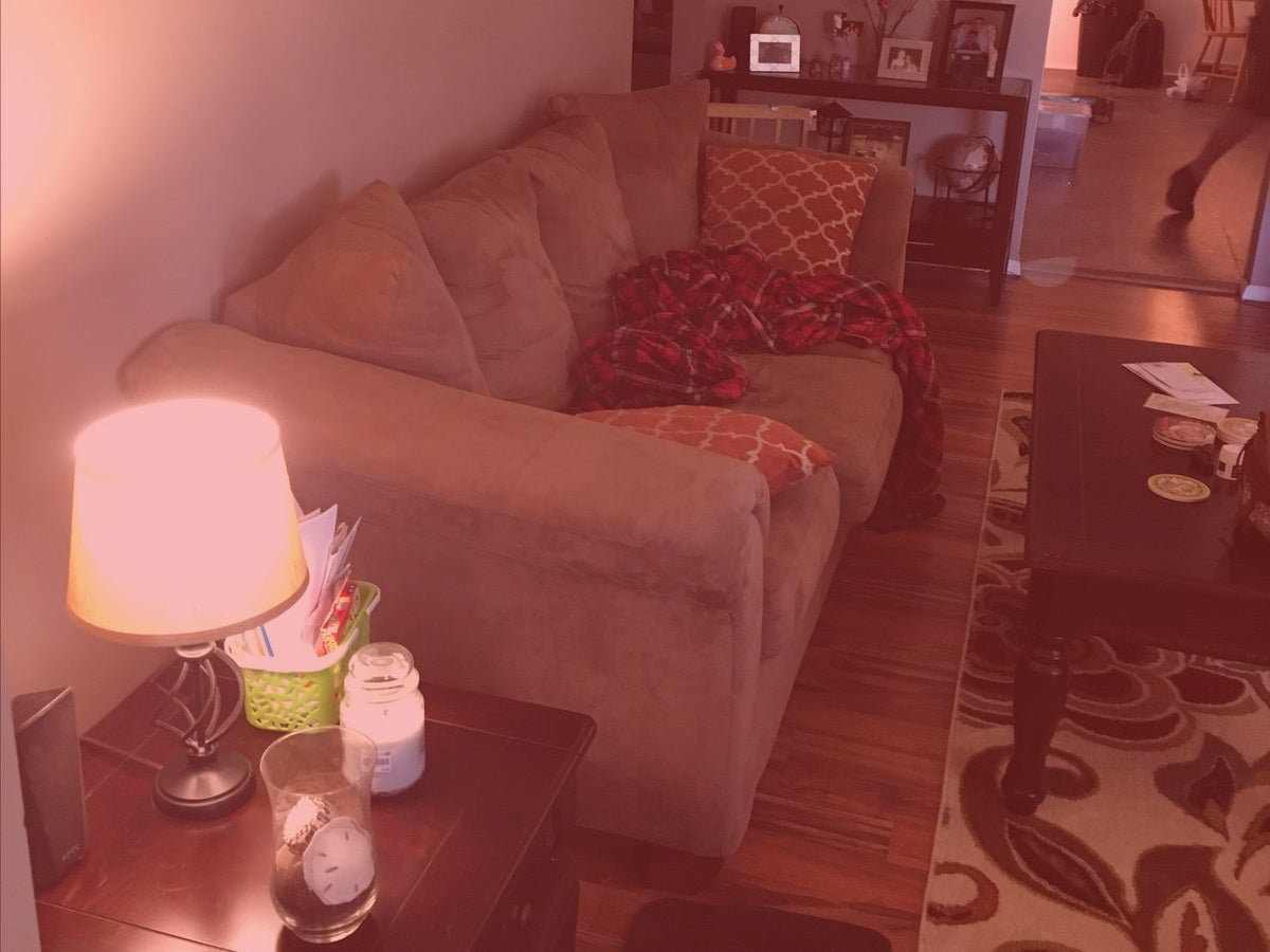 A living room photographed too close. A beige sofa fills the frame with its right arm cut off, a lamp and a cluttered side table crowd the foreground, and almost no bare floor is visible.