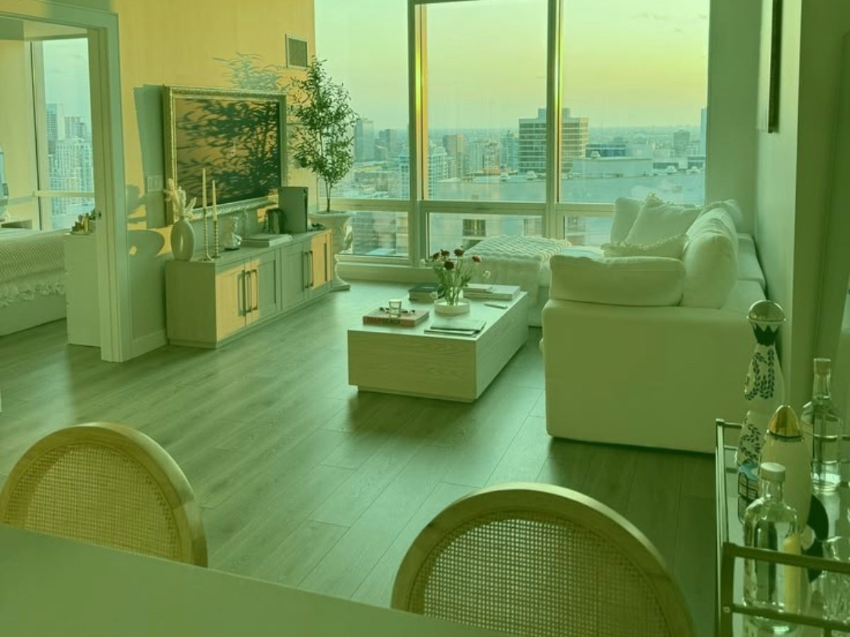 An open-plan living room photographed from far enough back. The whole sofa is in frame, a wide stretch of bare pale floor runs across the front, and daylight comes from a wall of windows.
